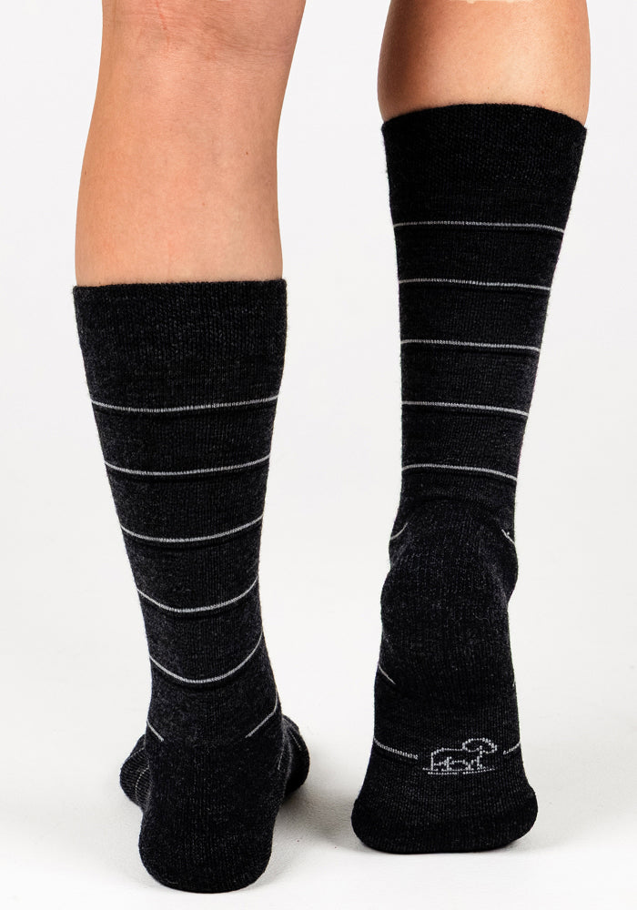 A person models the Woolx Womens Chandler Mid Calf Sock in dark grey charcoal, featuring thin white stripes, standing on a white background with the back of their legs and feet visible.