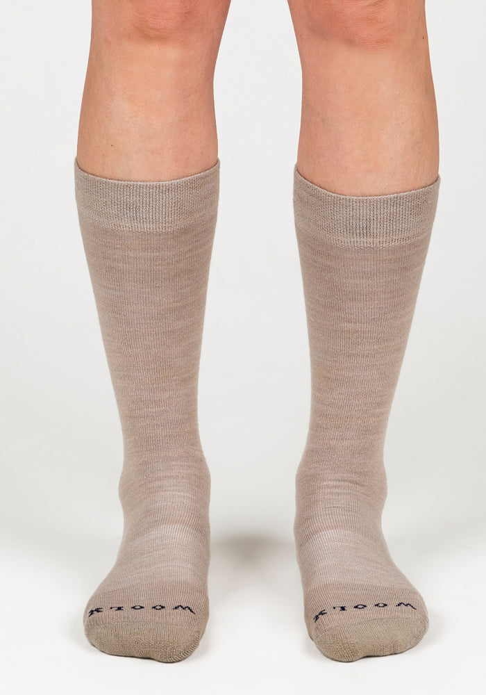 A pair of feet wear Woolx Womens Chandler Mid Calf Socks in Simply Taupe, standing on a white surface with toes forward. The lightweight Merino wool socks feature small text near the toes.