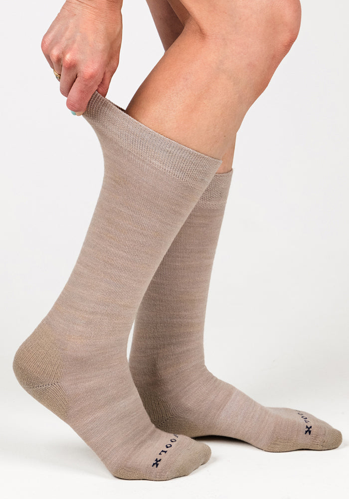 A person pulls up a pair of Woolx Womens Chandler Mid Calf Socks in Simply Taupe, holding the top with one hand. The foot features small text on the toes, and the background is plain white.