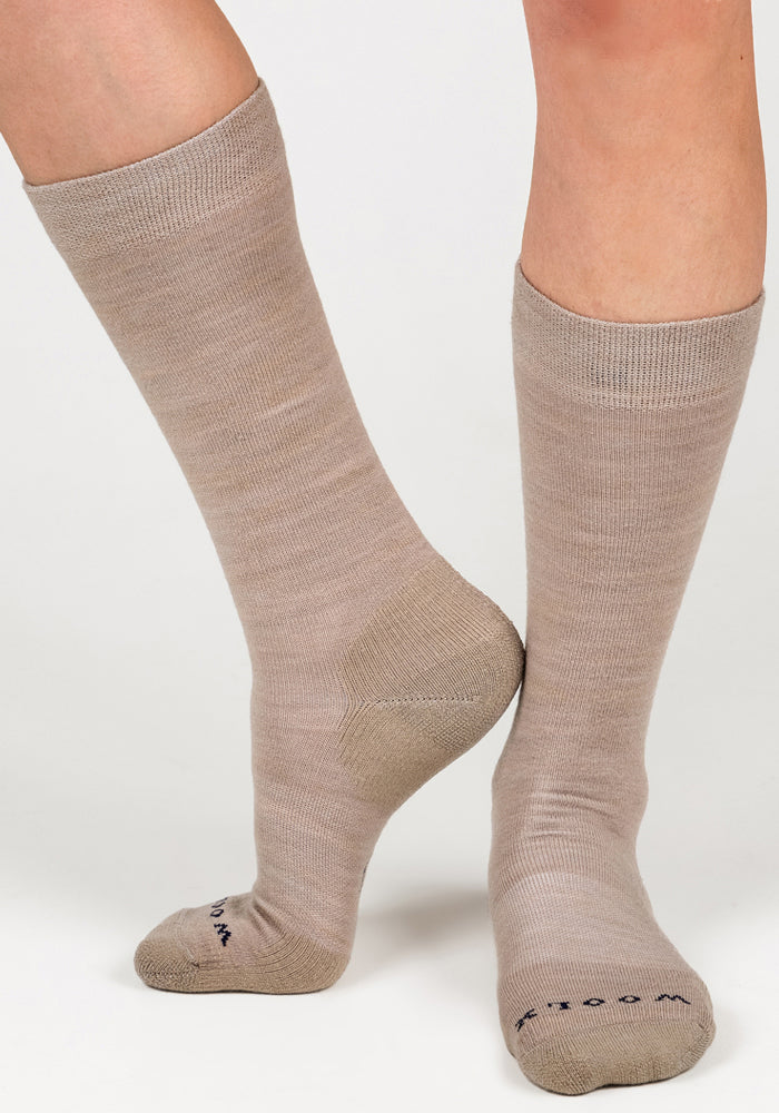 A person wears Woolx Womens Chandler Mid Calf Socks in Simply Taupe, standing with one foot flat and the other heel raised. The socks feature subtle toe text and are shown against a plain white background.