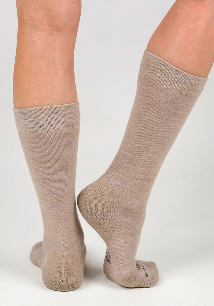 A pair of legs wear Woolx Womens Chandler Mid Calf Socks in Simply Taupe, set against a white background. One foot rests flat while the other lifts to display the heel and sole of these Merino wool socks.