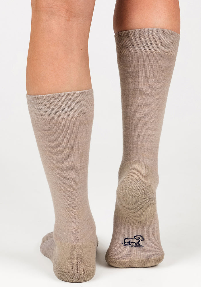 A close-up of two legs wearing Woolx Womens Chandler Mid Calf Socks in Simply Taupe. One sock features a small dark blue animal logo near the heel. The background is plain white.