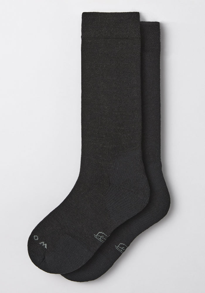 The Woolx Womens Chandler Mid Calf Sock in black is pictured laid flat on a light gray background, with one sock overlapping the other. It features a simple, minimal design and subtle toe text.