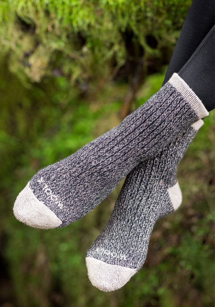 A woman wears Woolx Womens Cozy Mini Crew Socks in black, featuring thick knit Merino wool with cream-colored toes, heels, and cuffs; her legs are crossed outdoors against a blurred green background.