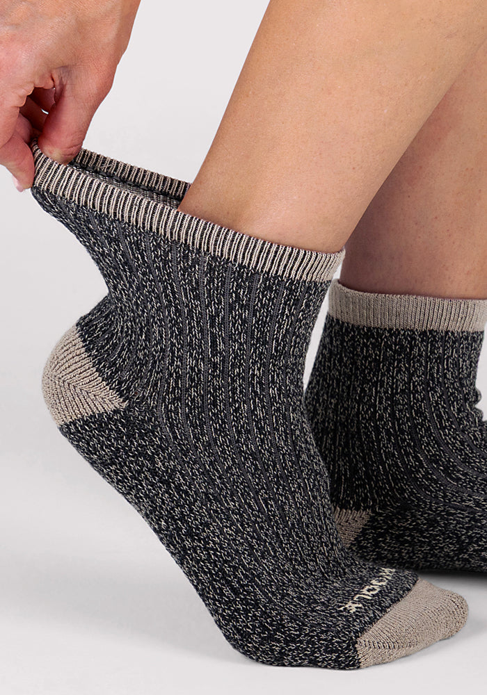 A close-up of someone pulling on a Woolx Womens Cozy Mini Crew Sock in ribbed dark gray and beige Merino wool, with the other foot wearing a matching sock, set against a light background.