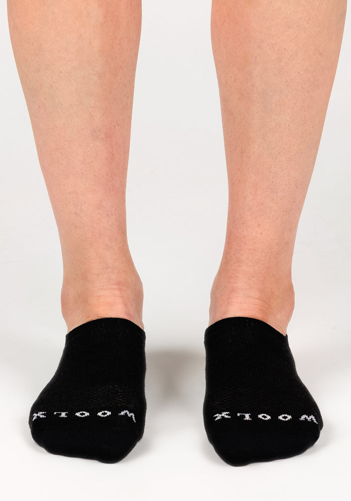 A person wearing Woolx Womens Daisy Ped Socks - Black Pepper, ultralight Merino wool minimalist socks, stands on a white surface. "WOOLX" is printed in white near the toes on each sock.