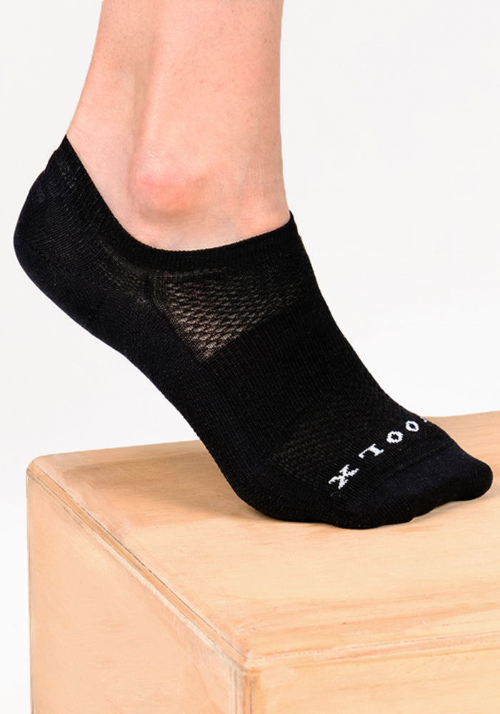 A person wears Woolx Womens Daisy Ped Socks in Black Pepper, featuring white text, while standing on a light wooden platform against a plain white background—ideal for minimalist sock enthusiasts.