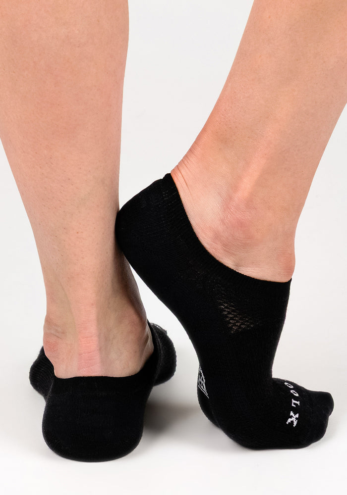 A close-up of two feet wearing Woolx Womens Daisy Ped Socks in Black Pepper, one foot flat and the other raised with toes pointed down, set against a plain white background.