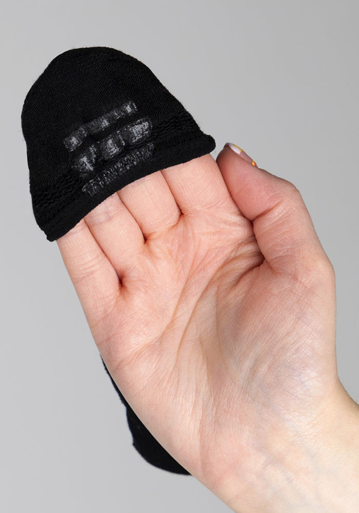 A hand holds a Woolx Womens Daisy Ped Sock in Black Pepper, turned inside out to reveal the non-slip grip pattern on the heel. The background is plain and light-colored.
