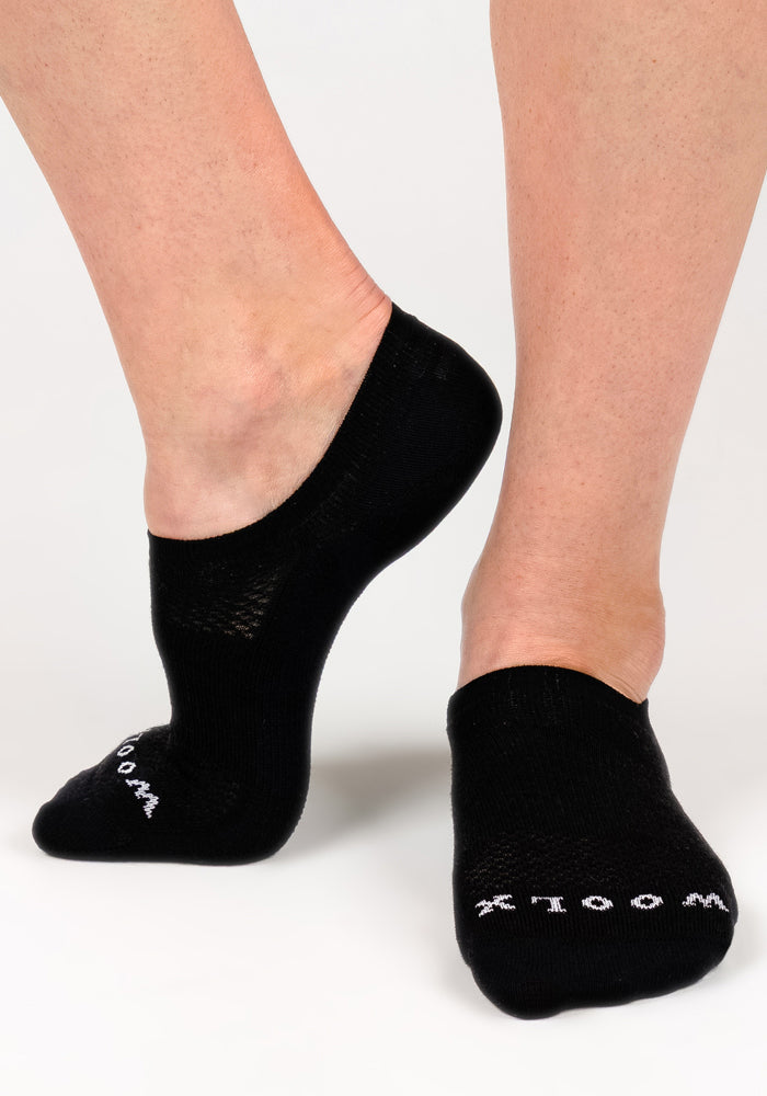 A close-up shows two feet wearing Woolx Womens Daisy Ped Socks in Black Pepper, minimalist Merino wool socks with "WOOL" in white text near the toes, set against a plain white background.