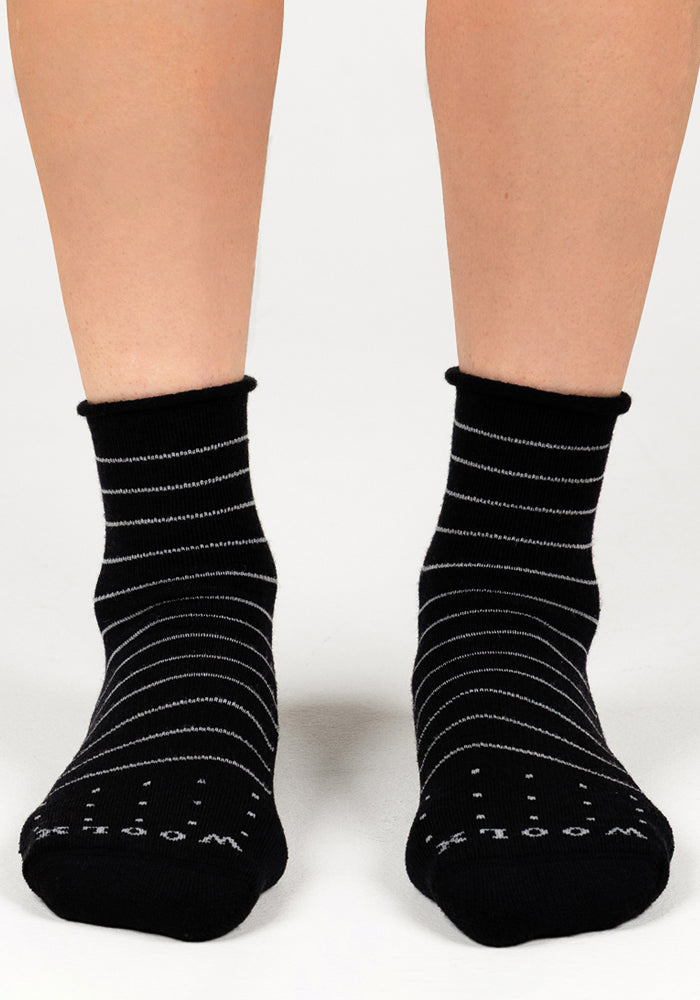 A pair of feet wear Woolx Womens Derby Roll Top Socks in black, featuring thin white stripes and "Monday" in white text on the toes. The breathable, comfortable socks are shown on a light surface.