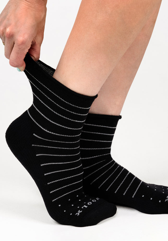 A person pulls up a Woolx Womens Derby Roll Top Sock in black with thin white stripes while the other foot already has the matching sock on. The breathable socks stand out against a plain white background.