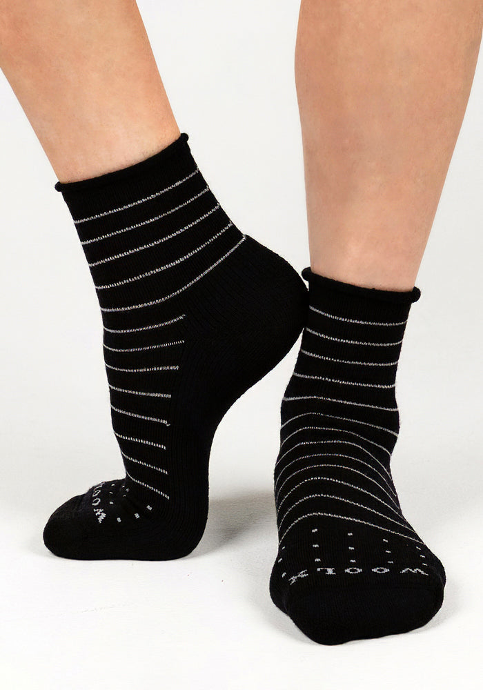 A pair of feet wearing Woolx Womens Derby Roll Top Sock in black, featuring breathable fabric, thin white stripes, and non-slip dots on the soles for extra grip, displayed against a plain white background.