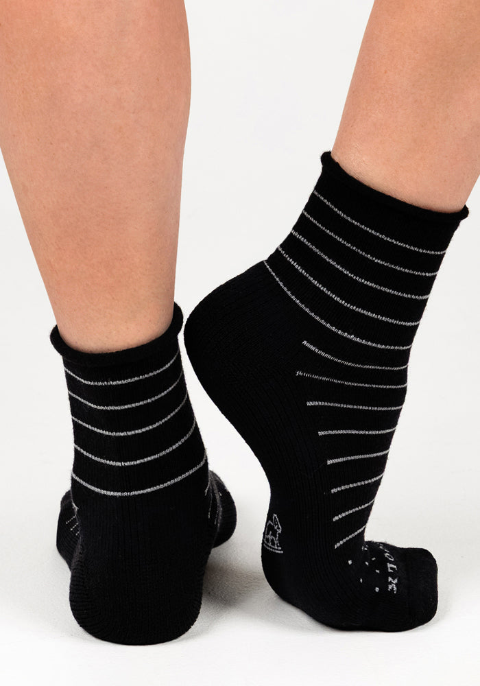 A pair of feet wear the Woolx Womens Derby Roll Top Sock in black, featuring thin white stripes. Shown from the back with raised heels, these stylish and breathable socks are highlighted against a plain white background.