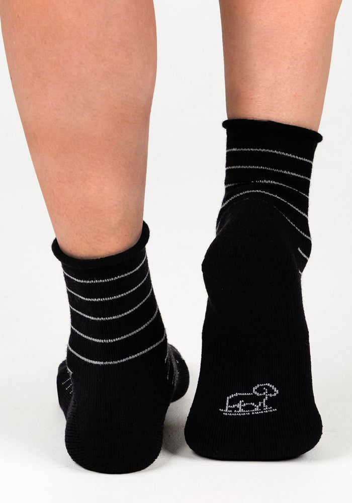 A close-up of two feet in Woolx Womens Derby Roll Top Socks - Black: the left sock has thin white stripes, and the right displays a small white dog outline near the heel, all against a crisp white background.