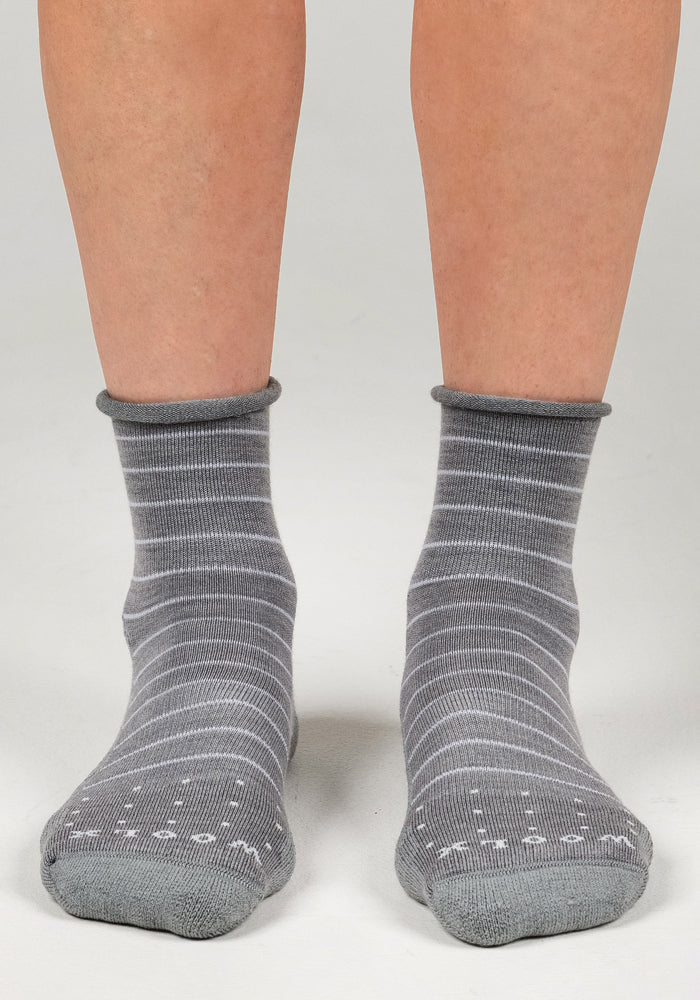 A pair of feet wearing Woolx Womens Derby Roll Top Sock - Grey with white stripes and grip dots on the soles, standing on a light gray surface. These breathable socks feature a stylish rolled cuff.