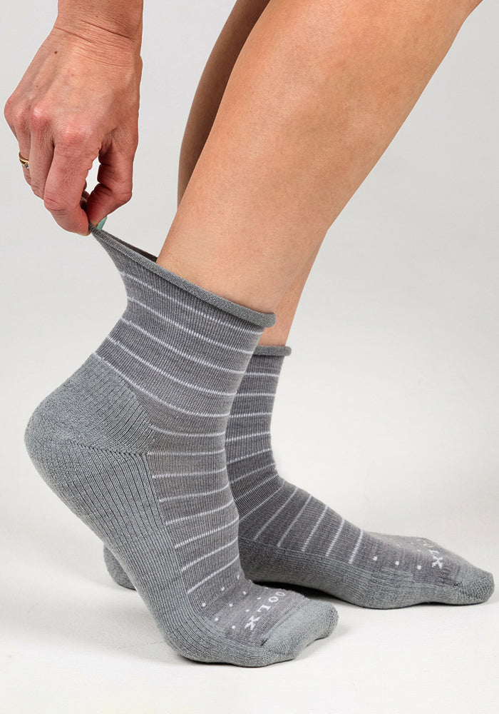 A person pulls up a Woolx Womens Derby Roll Top Sock - Grey with white stripes on their left foot, while the matching breathable sock is already on their right. The background is plain and light-colored.