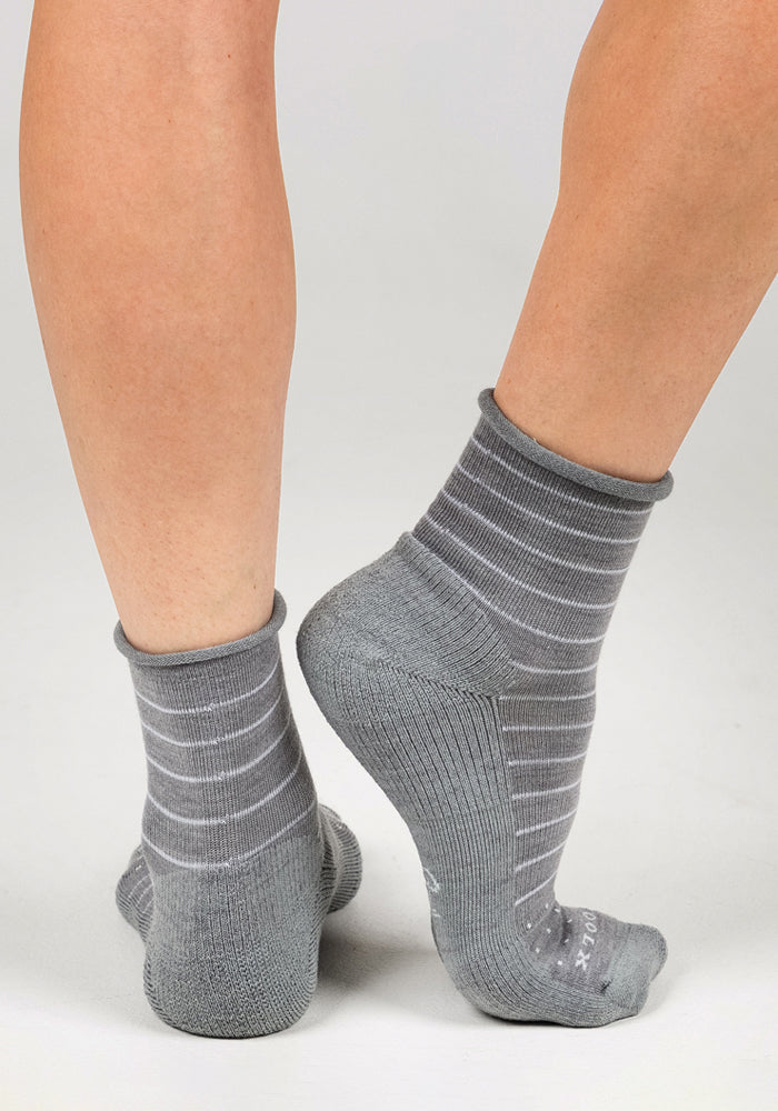 A person stands on tiptoe, displaying the back of their legs and feet in Woolx Womens Derby Roll Top Sock - Grey, featuring white horizontal stripes, against a plain background.