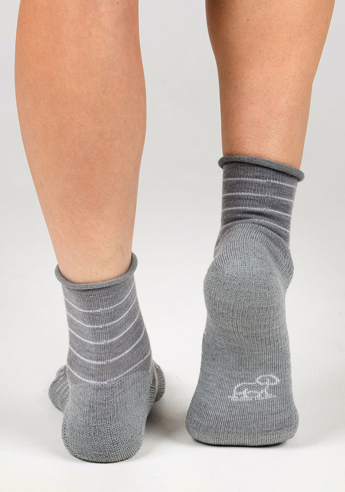 A person is shown from behind wearing the Woolx Womens Derby Roll Top Sock in grey, featuring white stripes, a logo on the heel, breathable fabric, and ankle-length coverage, standing on a white surface.