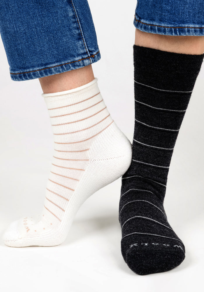 A person wears mismatched socks: a Woolx Womens Derby Roll Top Sock in Sand Dollar and a black sock with thin white stripes. Both breathable socks are styled with blue jeans featuring frayed hems.