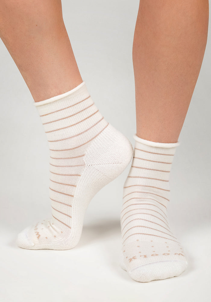 A person models the Woolx Womens Derby Roll Top Sock - Sand Dollar, showing both side and back views of the breathable white socks with thin beige stripes on a light surface.