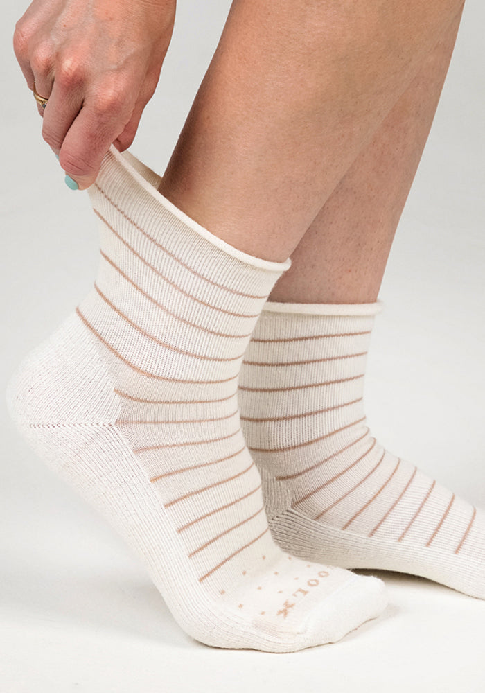 A person pulls on the Woolx Womens Derby Roll Top Sock - Sand Dollar, featuring thin, light brown stripes. The ankle-length sock is gently stretched at the top, showcasing its comfortable fit and breathable design.