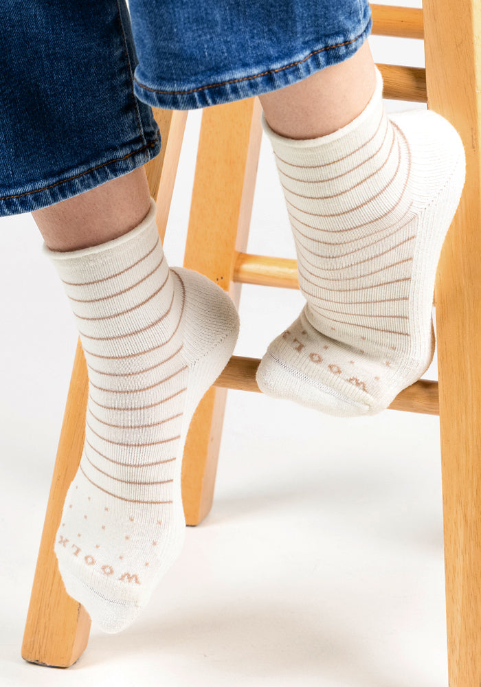 Someone wearing Woolx Womens Derby Roll Top Socks in Sand Dollar rests their feet on a light wooden stool's footrest. They are also wearing blue jeans.