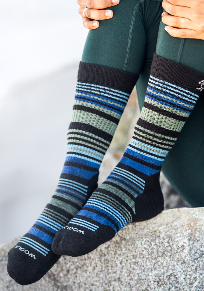 A person wearing dark green leggings and Woolx Womens Everyday Crew Socks in Black Stripe sits on a rock, hands resting on their knees.