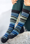 A person wearing dark green leggings and Woolx Womens Everyday Crew Socks in Black Stripe sits on a rock, hands resting on their knees.