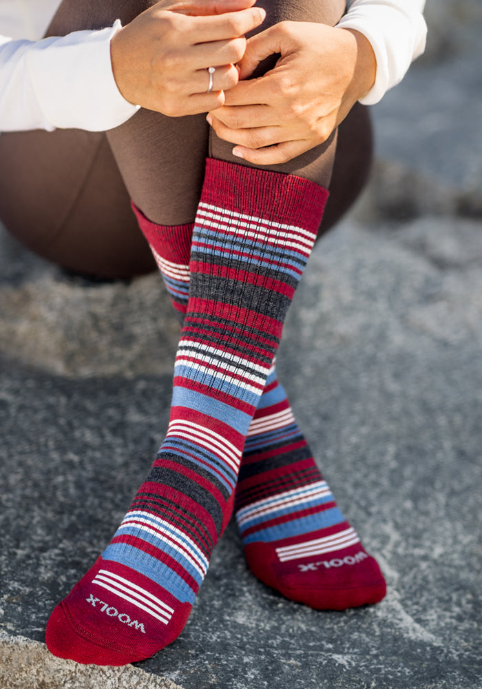 A person sits on a rock wearing Woolx Womens Everyday Crew Socks in Rhubarb Stripe—colorful, moisture-wicking Merino wool socks featuring red, blue, gray, and white stripes with "Woolx" branding on the toes. Their legs are crossed and hands rest on their knee.