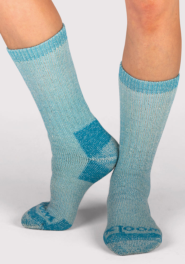 A person wearing Woolx Womens Extreme Crew Socks in Real Teal, featuring darker blue heels, toes, and toe lettering, stands on a light gray background with one foot flat and the other raised on the toes.