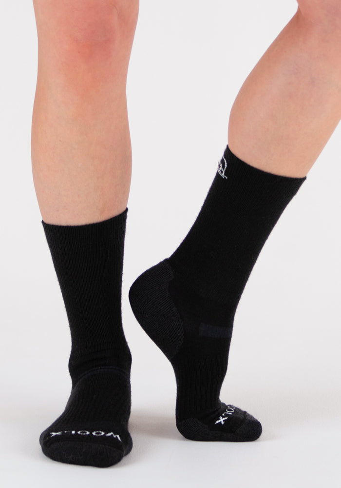 A person wears Woolx Harbor Crew Sock Light Cushion - Black, standing on a white background with one foot flat and the other raised. These socks regulate temperature and feature subtle logos near the toes and ankle.