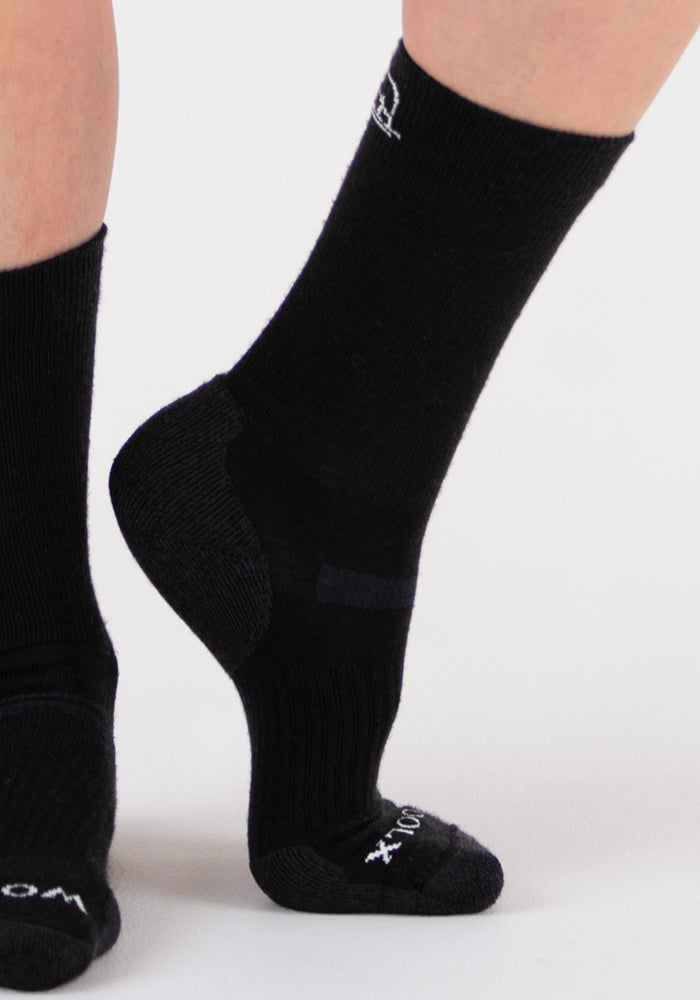 A close-up of two feet wearing Woolx Harbor Crew Sock Light Cushion in black on a light background—one foot raised to reveal the heel, and the other flat, showcasing their temperature-regulating and moisture-wicking comfort.