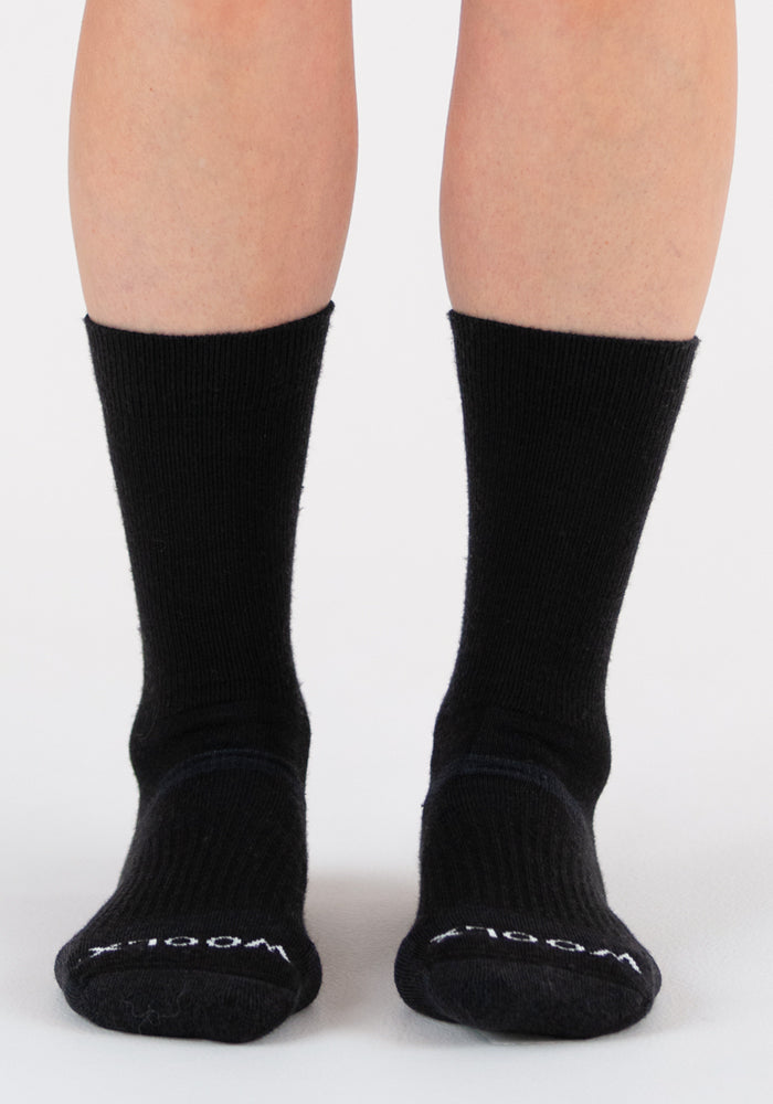 A person wears Woolx Harbor Crew Sock Light Cushion in black, standing on a white surface. The ribbed, moisture-wicking Merino wool socks show "WOOL" near the toes. Only the lower legs and feet are visible.