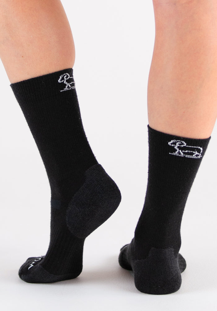 A person wearing Woolx Harbor Crew Sock Light Cushion in black, featuring a white dog outline on the back, stands on a white surface. These Merino wool socks regulate temperature, keeping feet comfortable in any condition.
