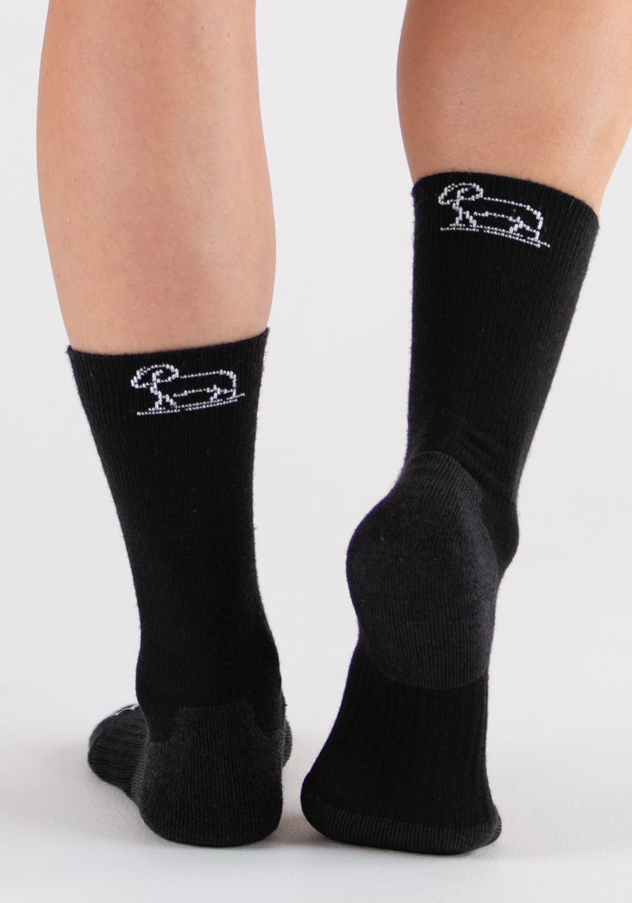 A person stands on a white surface with heels facing the camera, wearing black Woolx Harbor Crew Sock Light Cushion socks featuring a white rabbit outline on the back and made from temperature-regulating Merino wool.