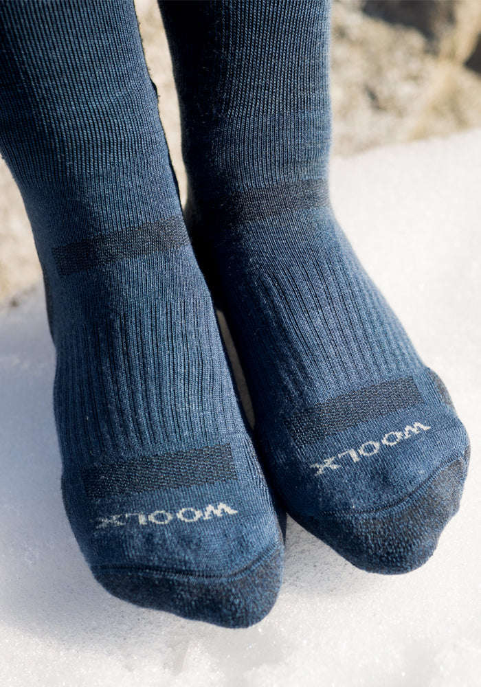 A close-up of two feet in Woolx Womens Harbor Crew Socks, dark blue with "WOOL" in white on the toes, standing on snow.