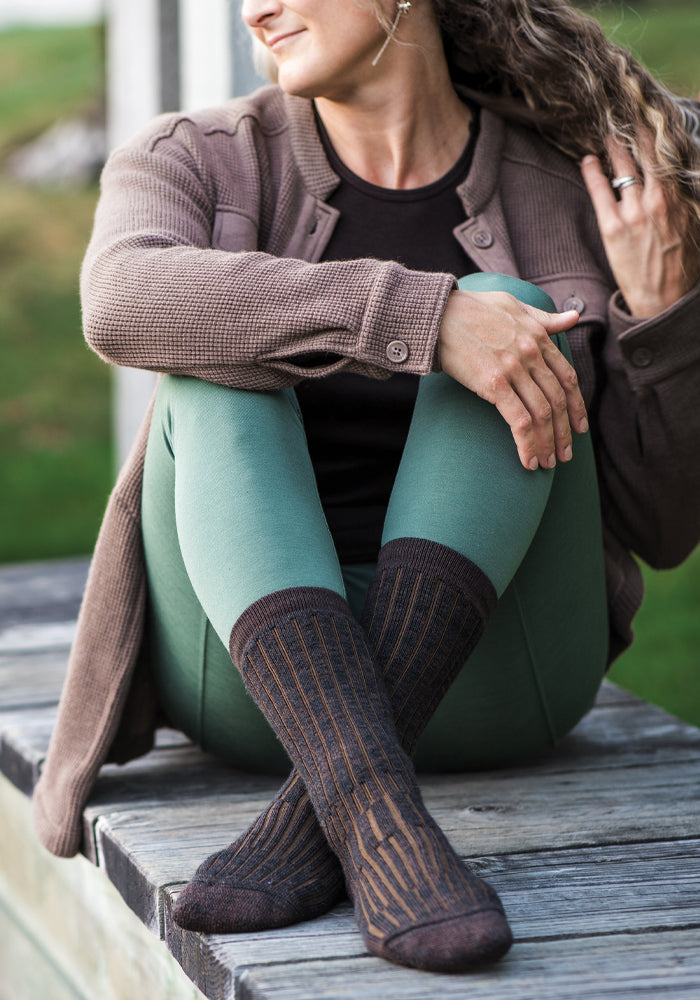 A person with curly hair sits outdoors on a wooden bench, head turned, wearing a brown jacket, green leggings, and Woolx Womens Joey Mid Calf Socks in Dark Espresso with a vertical striped pattern.