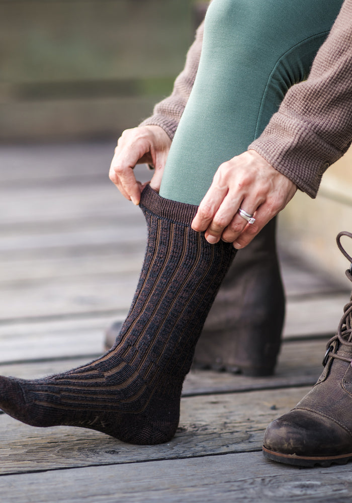 A person in green leggings and brown boots adjusts Woolx Womens Joey Mid Calf Socks in Dark Espresso while seated on a wooden surface.