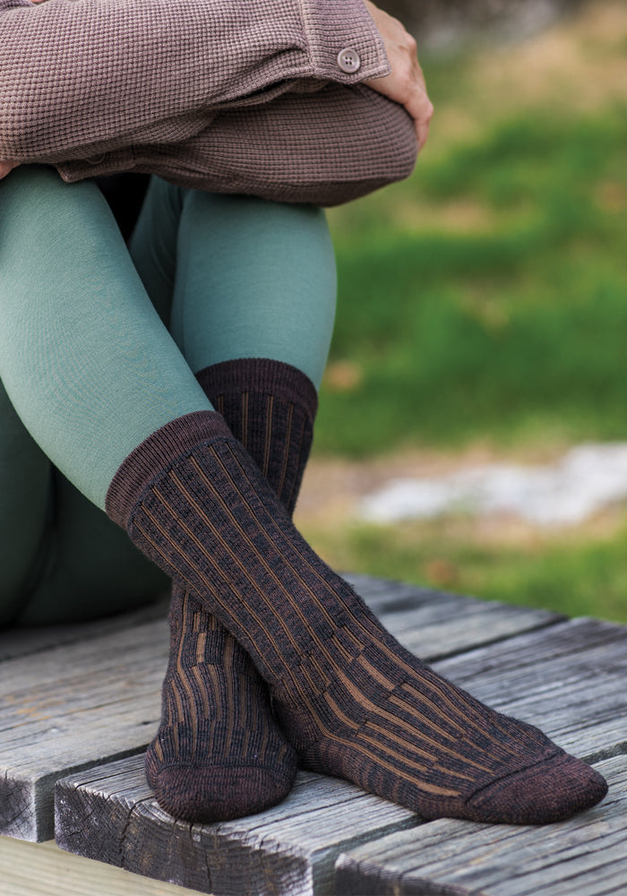 A person wearing mint green leggings, a brown textured jacket, and Woolx Womens Joey Mid Calf Socks in Dark Espresso sits cross-legged on a weathered wooden bench outdoors, with green grass in the background.