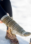 Wearing Woolx Womens Joey Mid Calf Socks in Dark Forest Marled and brown lace-up boots, a person sits on snow, pulling up one sock over black leggings.