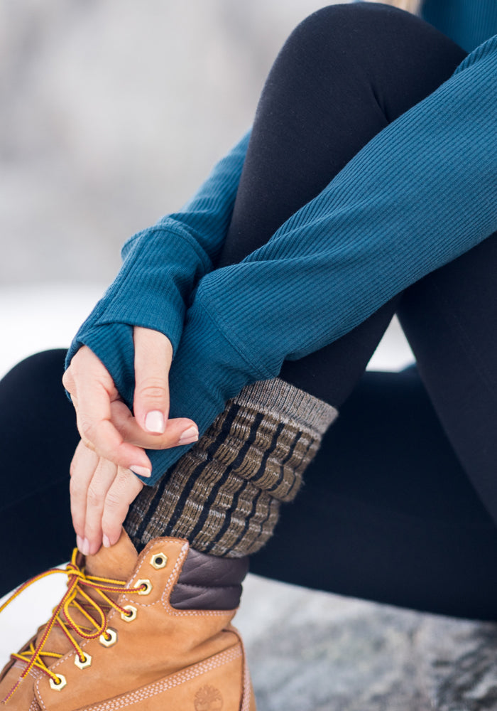 A person wearing black leggings, a blue long-sleeve shirt, Woolx Womens Joey Mid Calf Socks, and tan lace-up boots sits outdoors, resting their hands on their leg.