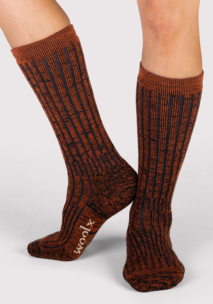 A person wears Woolx Womens Joey Mid Calf Socks in Cinnamon Spice, featuring a ribbed, heathered orange and dark gray pattern. These cozy Merino wool socks offer moisture-wicking comfort and display "ROOTS" on one sole.