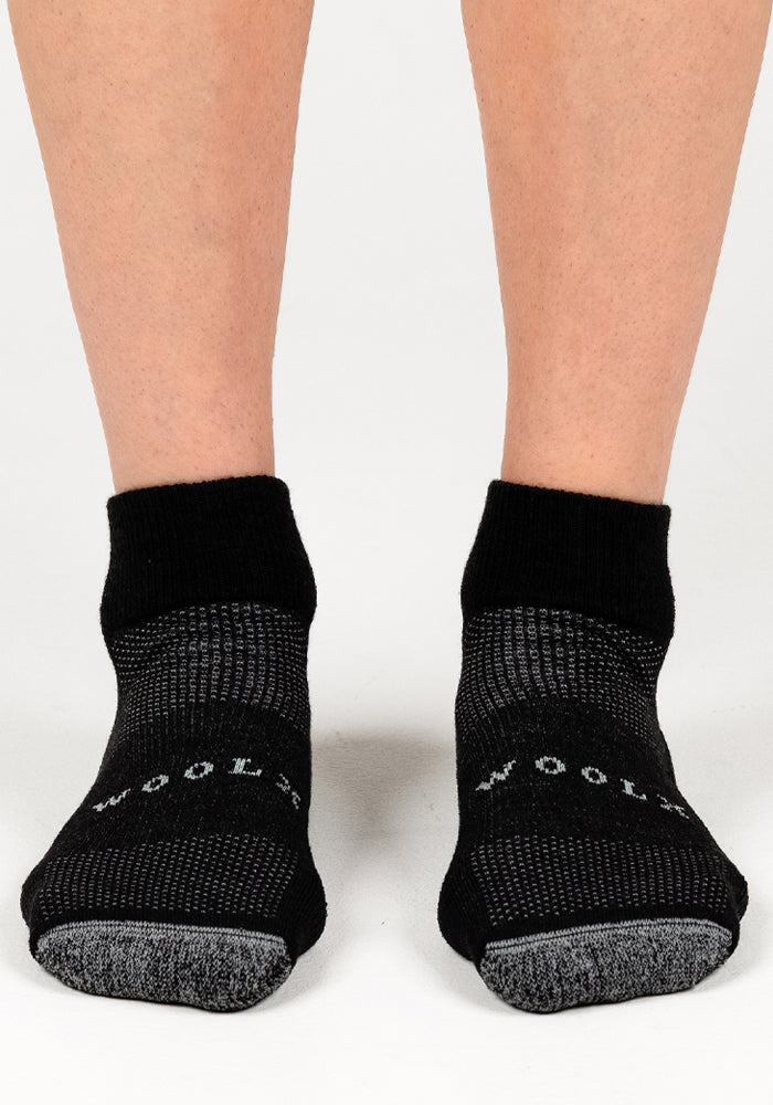 A pair of feet in black and gray Woolx Womens Rambler Ankle Socks - Black Pepper, with "WOOL" partially visible on the toes, stand on a white surface—ideal for comfort and moisture management.
