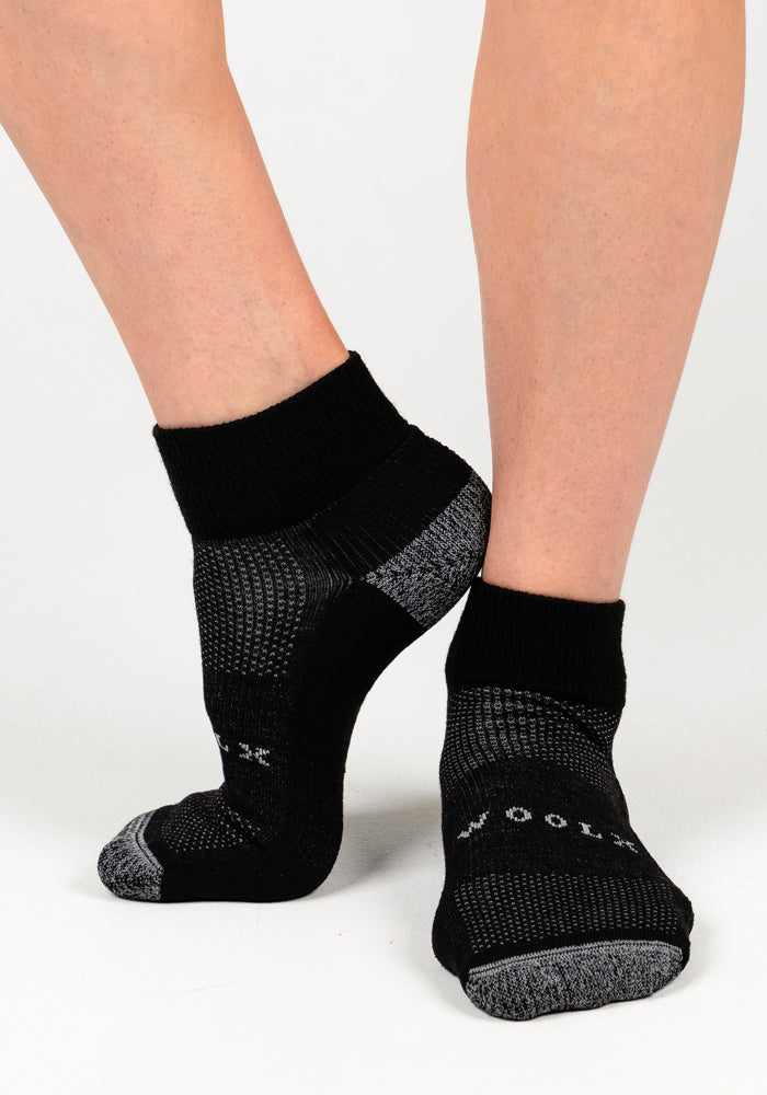 A person wearing Woolx Womens Rambler Ankle Sock in Black Pepper stands on a white background, one foot behind the other. "WOOLX" appears on the sole, showcasing these ankle socks’ excellent moisture management.