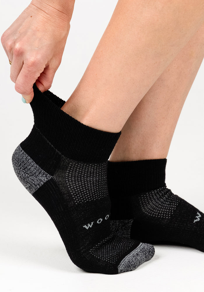 A person pulls on a Woolx Womens Rambler Ankle Sock in Black Pepper, adjusting the top for comfort and moisture control, while their other foot displays the matching Merino wool sock against a white background.