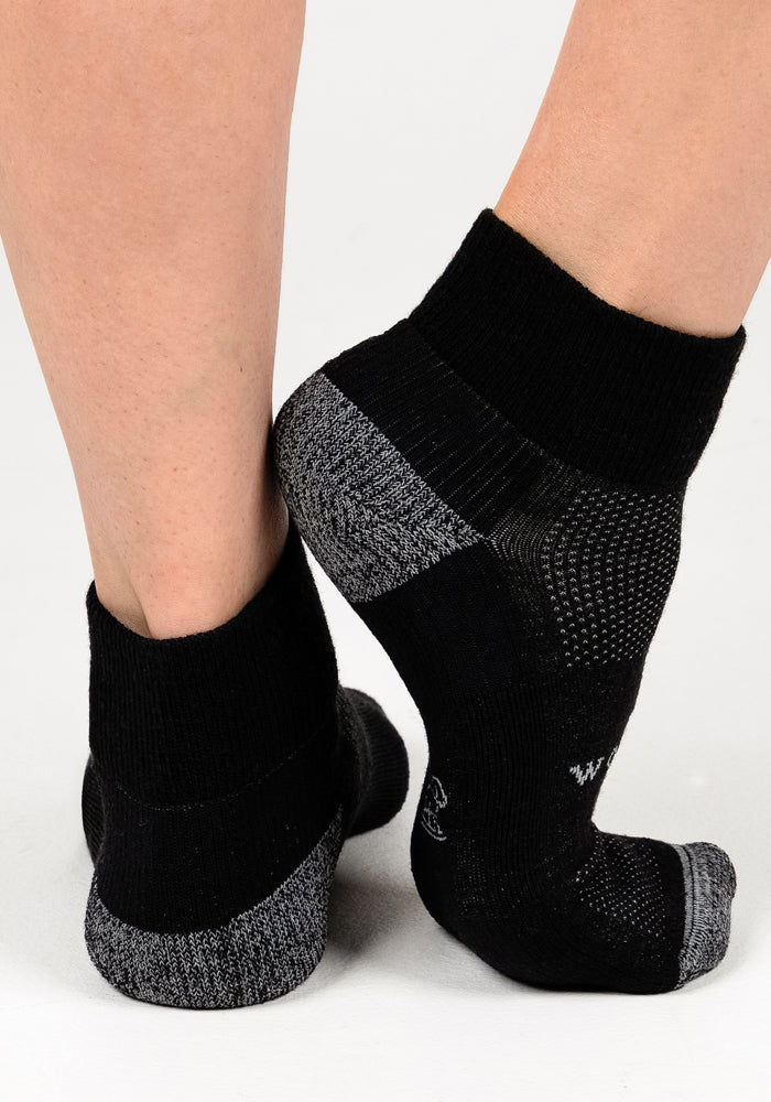 Two feet wear Woolx Womens Rambler Ankle Sock - Black Pepper, standing on a white surface—one foot on tiptoe and one flat—to showcase the comfort and moisture-wicking Merino wool design.