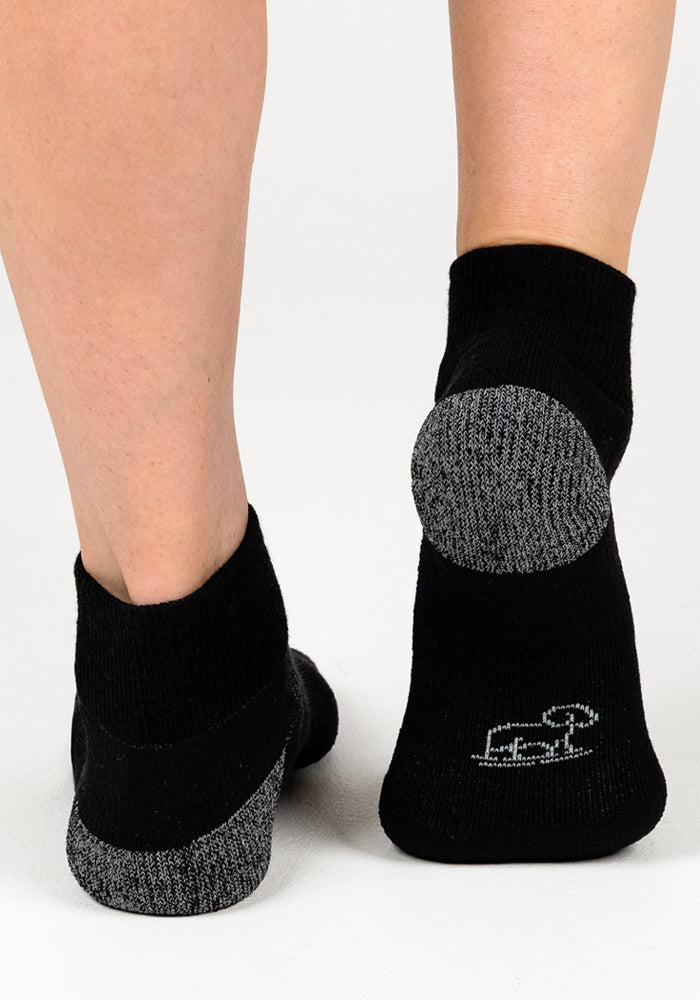 Close-up of two feet in Woolx Womens Rambler Ankle Sock - Black Pepper, with gray reinforced heels/toes and a small white logo near the sole. Shown on a white surface to highlight its excellent moisture management.