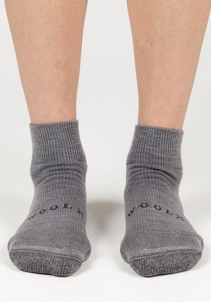 A person wearing Woolx Womens Rambler Ankle Sock - Pebble stands on a white background. Only the lower legs and feet are visible, and "WOOLX" is labeled near the toes. These socks offer excellent moisture management.