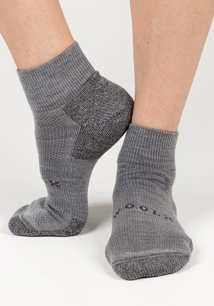 A close-up of two feet in Woolx Womens Rambler Ankle Sock - Pebble shows one foot flat, the other on its toes, highlighting the textured knit, ribbed cuffs, and excellent moisture management.